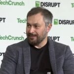 A bearded man grins against a TechCrunch-branded backdrop