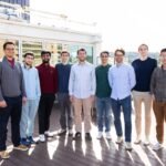 A group of nine men stand on a sunlit balcony.