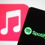 The Spotify logo seen displayed on a smartphone screen in front of Apple Music logo in the background.