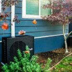 A dark grey heat pump sits outside a blue home.