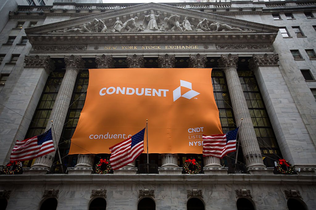 Conduent Inc. signage is displayed as American flags fly outside of the New York Stock Exchange (NYSE) in New York, U.S., on Tuesday, Jan. 3, 2017. U.S. stocks rose Tuesday, pointing to the biggest gain in a month for Wall Street's first session of the new year, helped by a string of robust economic data from China and Europe. Photographer: Michael Nagle/Bloomberg via Getty Images