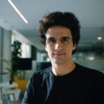 A man wearing a large black t-shirt stands in a sunlit office.