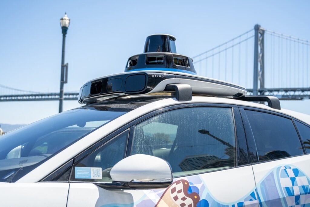 Self-driving Waymo car with rooftop lidar and bird-themed mural on the Embarcadero with the San Francisco-Oakland Bay Bridge in background, San Francisco, California, August 14, 2025.