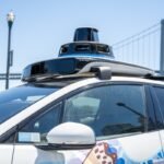 Self-driving Waymo car with rooftop lidar and bird-themed mural on the Embarcadero with the San Francisco-Oakland Bay Bridge in background, San Francisco, California, August 14, 2025.