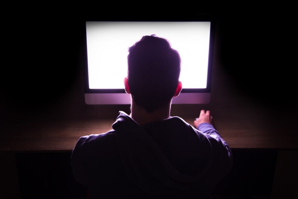 Back view of a man in front of bright computer working in the dark.