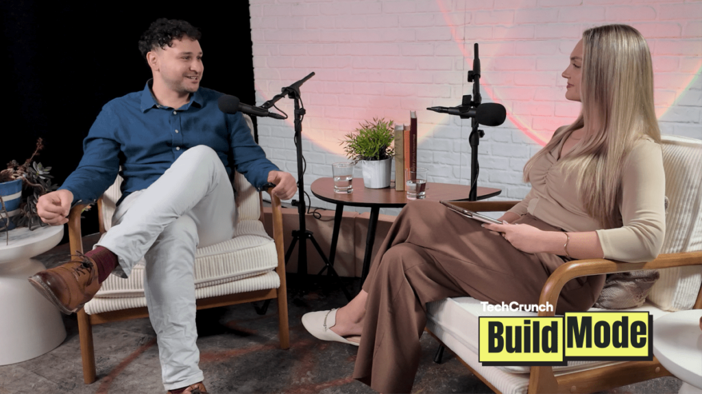 Two people seated in armchairs during a podcast interview, speaking into microphones across a small table with water glasses and a plant, with a “TechCrunch Build Mode” logo overlay in the corner.