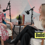 Two women sit facing each other in a podcast studio, speaking into microphones. One wears a red floral dress and glasses, while the other wears a cream top and black pants. A small table between them holds a bottle of wine, glasses of water, and books. A TechCrunch Build Mode logo appears in the corner.