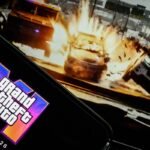 LONDON, ENGLAND - MAY 06: In this photo illustration, a phone displaying the Grant Theft Auto VI logo and new release date is shown in front of a screen displaying the second official trailer for the game, on May 06, 2025 in London, England. Last week, Rockstar Games, developer of the Grand Theft Auto franchise, announced the game had been pushed back to May of next year.