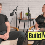 Artisan CEO, Jaspar Carmichael-Jack and Build Mode host Isabelle Johannessen sit facing each other in a podcast studio with microphones, mid-conversation, in front of a white brick wall; a small table with books and a plant sits between them, and a “TechCrunch Build Mode” logo overlay appears in the lower right corner.