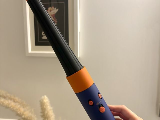 Mão segurando o MultiStyler e Secador Dyson Airwrap id, um modelador de cachos com corpo preto, alça azul e laranja e pequenos botões na alça