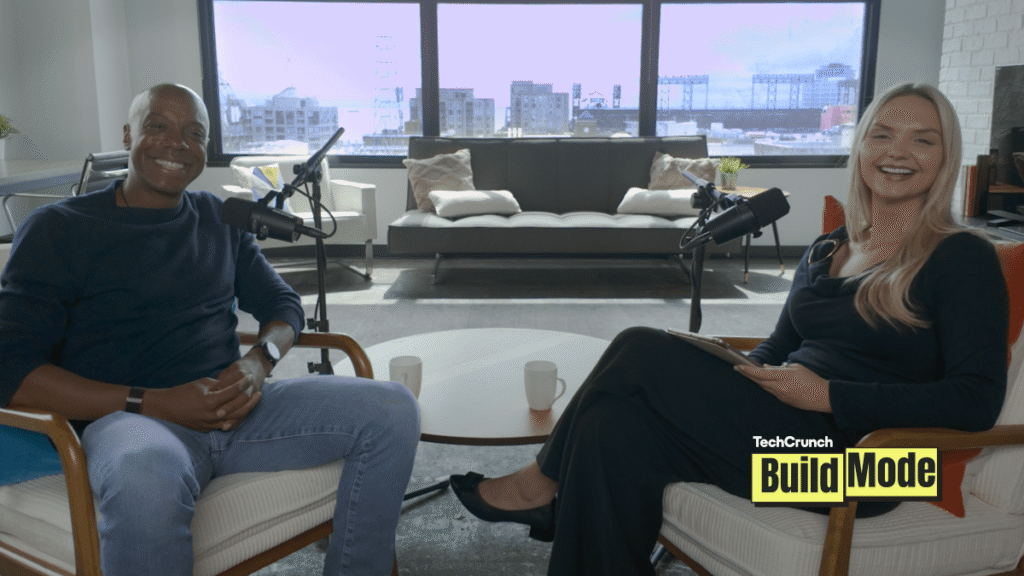 TechCrunch Build Mode podcast host Isabelle Johannessen sits across from venture capitalist Charles Hudson in a bright podcast studio. Both are smiling toward the camera, seated in armchairs with microphones positioned between them. A coffee table with two mugs sits in the foreground, while large windows behind them overlook the San Francisco skyline. The TechCrunch Build Mode logo appears in the bottom-right corner.