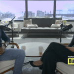 TechCrunch Build Mode podcast host Isabelle Johannessen sits across from venture capitalist Charles Hudson in a bright podcast studio. Both are smiling toward the camera, seated in armchairs with microphones positioned between them. A coffee table with two mugs sits in the foreground, while large windows behind them overlook the San Francisco skyline. The TechCrunch Build Mode logo appears in the bottom-right corner.